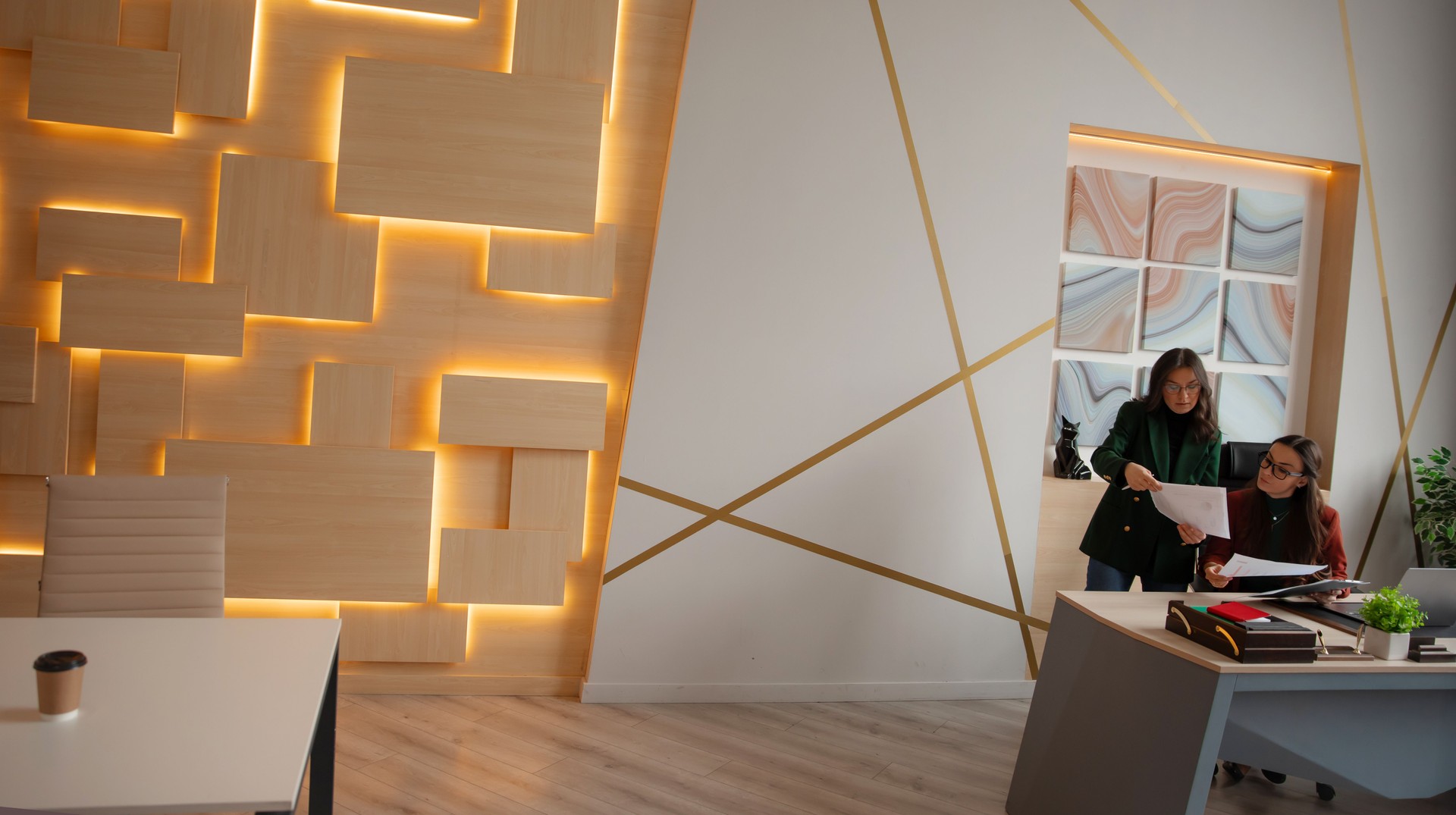 Two businesswomen in formal wear review documents at a modern office desk. Stylish workspace features abstract wall art, warm lighting, and a contemporary interior design.